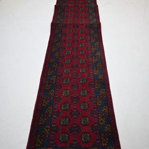 May include: A long, rectangular red rug with a repeating geometric pattern in navy blue, gold, and dark green. The rug has a central field of red with a series of medallions. The border features a complex design. The rug is displayed on a white surface.