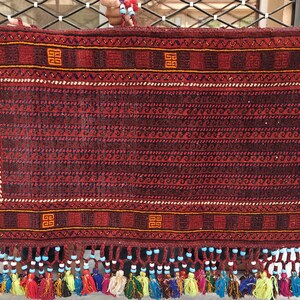 May include: A rectangular woven textile featuring a deep red and black geometric pattern with orange square motifs. The bottom edge is adorned with a row of light blue beads and a fringe of multi-colored yarn tassels in yellow, red, and green.