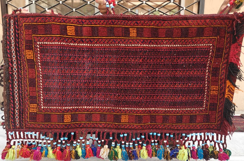 May include: A rectangular woven textile featuring a deep red and black geometric pattern with orange square motifs. The bottom edge is adorned with a row of light blue beads and a fringe of multi-colored yarn tassels in yellow, red, and green.