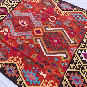 Op de afbeelding: Een geweven kleed met een levendig rood centraal veld met ingewikkelde geometrische patronen in tinten turquoise, magenta, geel en wit. Het kleed is omlijst door brede donkerbruine randen versierd met herhalende cr&egrave;mekleurige en zwarte hoekige motieven.