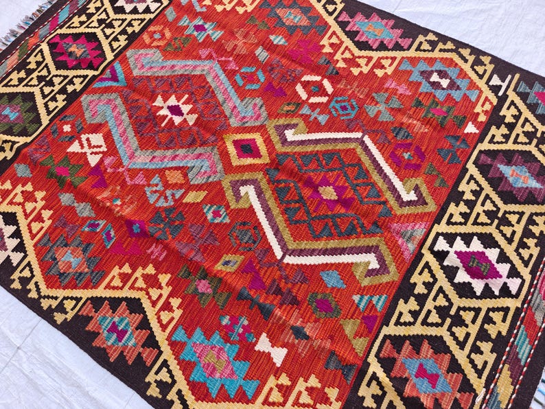 Op de afbeelding: Een geweven kleed met een levendig rood centraal veld met ingewikkelde geometrische patronen in tinten turquoise, magenta, geel en wit. Het kleed is omlijst door brede donkerbruine randen versierd met herhalende cr&egrave;mekleurige en zwarte hoekige motieven.