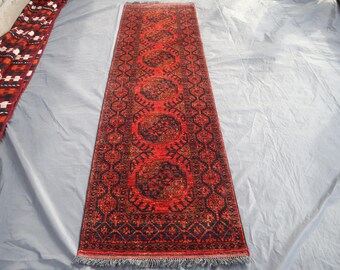 May include: A long, rectangular red rug with a detailed, repeating pattern in dark blue and gold. The rug features a central design of connected medallions and a fringed edge. The rug is displayed on a light gray surface.