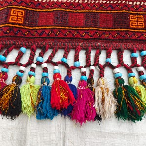 May include: A woven textile border featuring a deep red background with geometric orange and black patterns. The bottom edge is adorned with a row of braided fringe, each tipped with a colorful tassel and accented by bright turquoise and white beads.