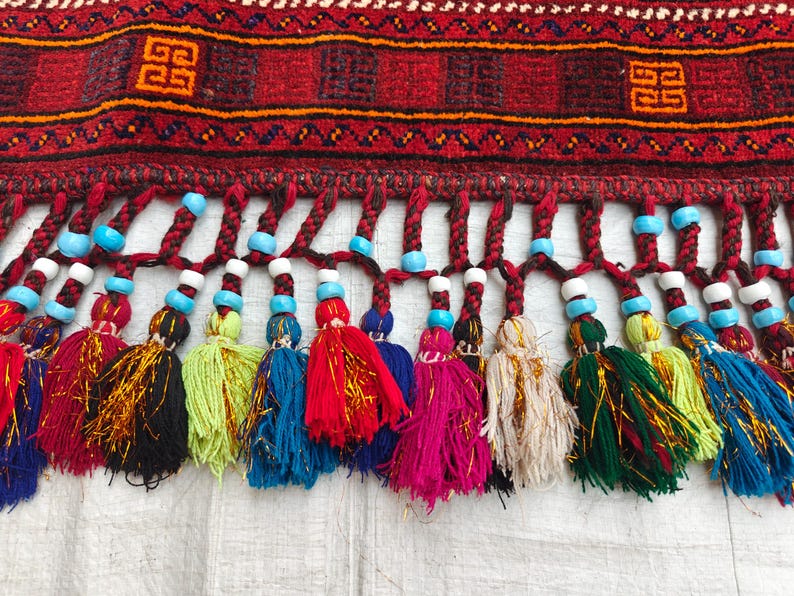 May include: A woven textile border featuring a deep red background with geometric orange and black patterns. The bottom edge is adorned with a row of braided fringe, each tipped with a colorful tassel and accented by bright turquoise and white beads.