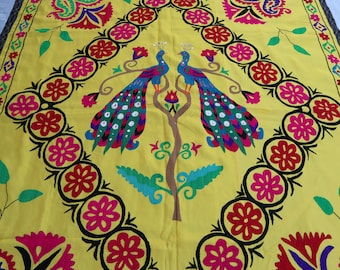 May include: A yellow fabric tapestry with a colourful embroidered design featuring two peacocks with their tails spread out. The peacocks are facing each other and are surrounded by floral motifs. The tapestry has a black border with a repeating diamond pattern.