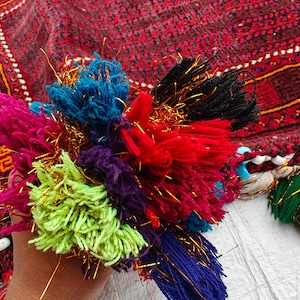 May include: A cluster of multi-colored yarn tassels in shades of lime green, magenta, blue, purple, red, and black, interwoven with metallic gold threads. The tassels are attached to a dark red patterned textile featuring light blue and white beads.