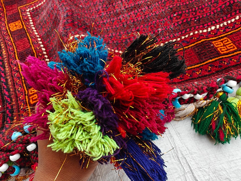 May include: A cluster of multi-colored yarn tassels in shades of lime green, magenta, blue, purple, red, and black, interwoven with metallic gold threads. The tassels are attached to a dark red patterned textile featuring light blue and white beads.