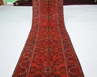 May include: A long, rectangular red rug with an intricate geometric pattern. The rug features a central design with repeating floral and geometric motifs in shades of red, brown, and black. The rug has a fringed edge.