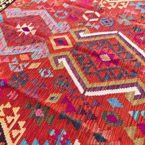 Op de afbeelding: Een geweven textielkleed met een levendige rode basiskleur en ingewikkelde geometrische patronen in tinten blauw, paars, wit en geel. Het ontwerp bevat herhalende ruit- en chevronvormen over het gehele oppervlak met een donkere rand.