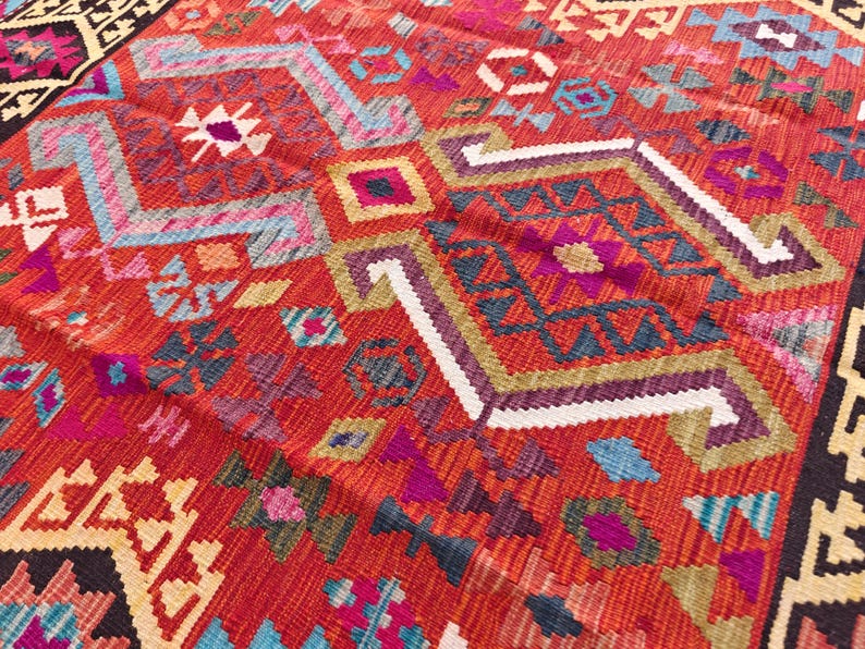 Op de afbeelding: Een geweven textielkleed met een levendige rode basiskleur en ingewikkelde geometrische patronen in tinten blauw, paars, wit en geel. Het ontwerp bevat herhalende ruit- en chevronvormen over het gehele oppervlak met een donkere rand.