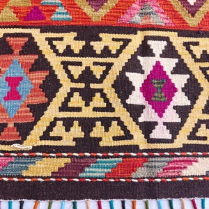 Op de afbeelding: Een geweven textielkleed met een herhalend geometrisch patroon in tinten donkerbruin, cr&egrave;me, felroze, lichtblauw en oranje. Het ontwerp bevat ruitvormige motieven en een horizontale rand met een veelkleurige franje aan de onderkant.
