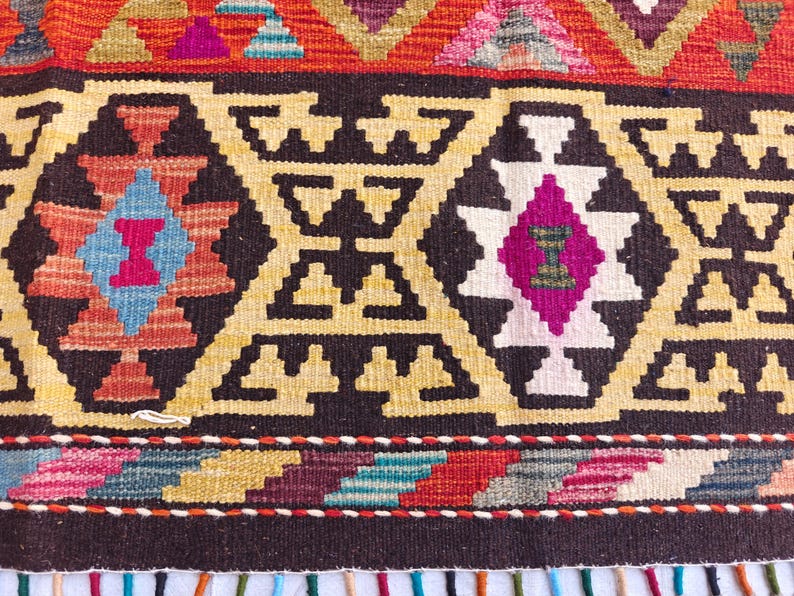Op de afbeelding: Een geweven textielkleed met een herhalend geometrisch patroon in tinten donkerbruin, cr&egrave;me, felroze, lichtblauw en oranje. Het ontwerp bevat ruitvormige motieven en een horizontale rand met een veelkleurige franje aan de onderkant.
