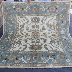 May include: A vintage rug with a cream background and a blue and brown floral pattern. The rug has a border with a repeating floral design.
