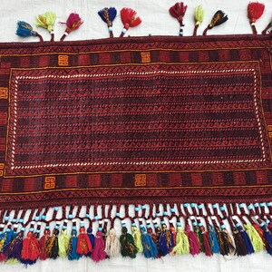 May include: A rectangular rug featuring a deep red and black geometric pattern with a central field of repeating wave motifs. The border includes orange square symbols. The edges are adorned with numerous multi-colored tassels and beads.