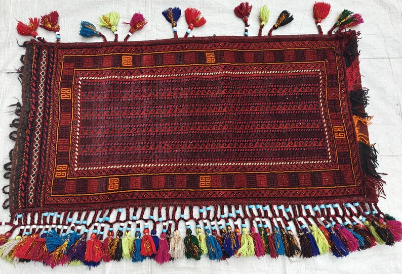 May include: A rectangular rug featuring a deep red and black geometric pattern with a central field of repeating wave motifs. The border includes orange square symbols. The edges are adorned with numerous multi-colored tassels and beads.