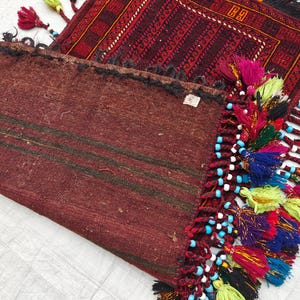 May include: A folded dark red textile with dark horizontal stripes and a decorative edge featuring multi-colored tassels in shades of yellow, blue, pink, and purple, accented with small light blue beads, layered over a patterned red rug.
