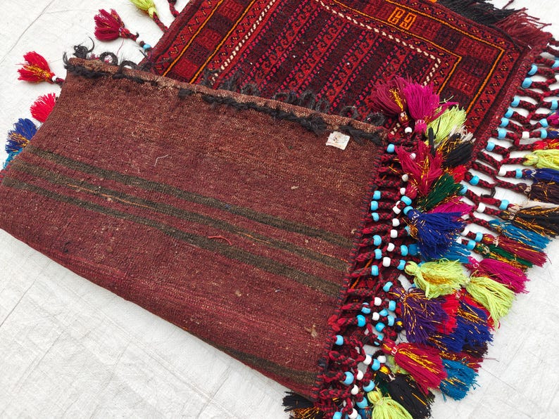 May include: A folded dark red textile with dark horizontal stripes and a decorative edge featuring multi-colored tassels in shades of yellow, blue, pink, and purple, accented with small light blue beads, layered over a patterned red rug.