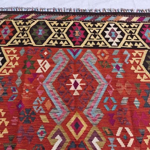 Op de afbeelding: Een geweven kleed met een levendige rode achtergrond en ingewikkelde geometrische patronen in donkerbruin, cr&egrave;me, lichtblauw, magenta en talingblauw. Het ontwerp bevat herhalende ruitvormen en hoekige motieven, afgewerkt met een franjerand.
