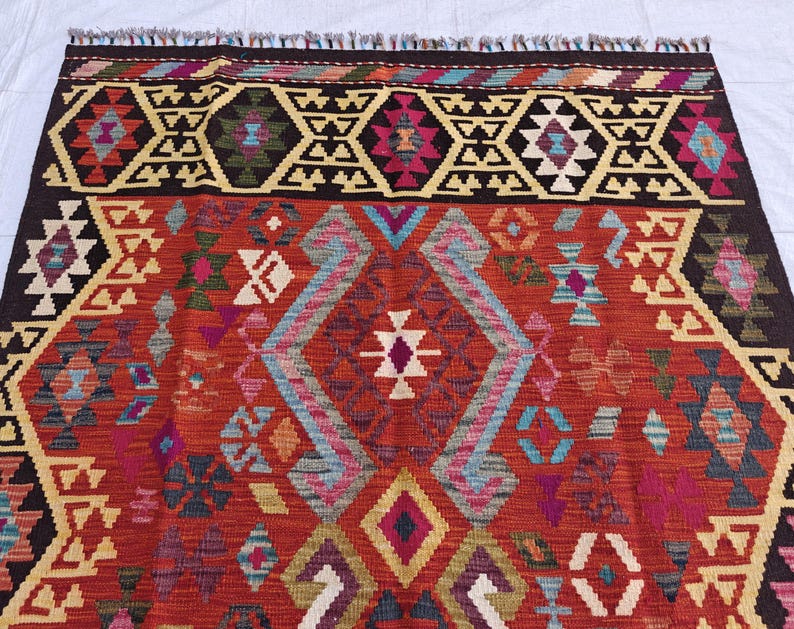 Op de afbeelding: Een geweven kleed met een levendige rode achtergrond en ingewikkelde geometrische patronen in donkerbruin, cr&egrave;me, lichtblauw, magenta en talingblauw. Het ontwerp bevat herhalende ruitvormen en hoekige motieven, afgewerkt met een franjerand.