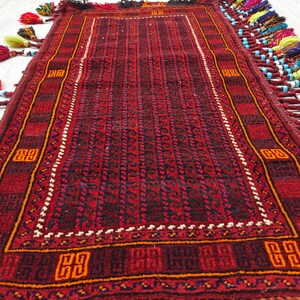May include: A rectangular rug featuring a deep red and black geometric pattern with a central field of repeating dark motifs. The border displays orange square accents, and the edges are adorned with long, multi-colored tassels and beads.