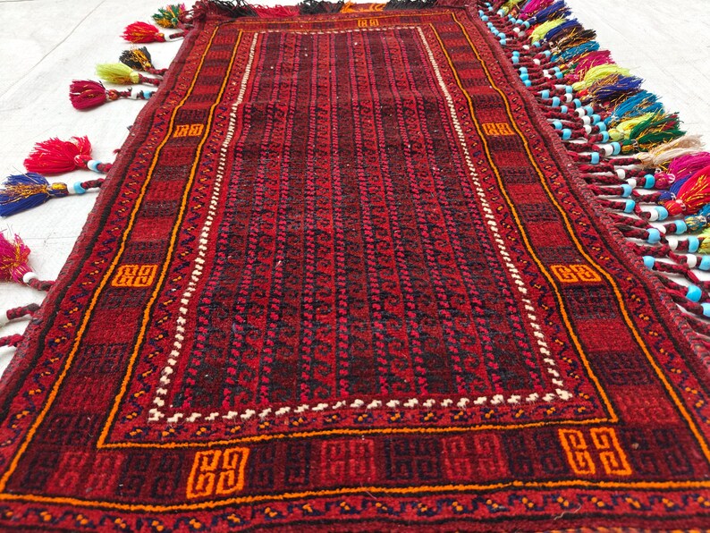 May include: A rectangular rug featuring a deep red and black geometric pattern with a central field of repeating dark motifs. The border displays orange square accents, and the edges are adorned with long, multi-colored tassels and beads.