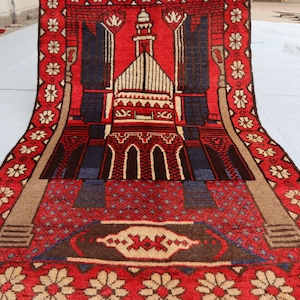 May include: A red prayer rug with a beige border and a detailed design of a mosque in the center. The rug has a floral pattern along the edges.
