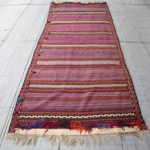 May include: A long, rectangular woven runner rug featuring horizontal stripes in shades of purple, magenta, and maroon. The rug includes intricate geometric patterns, small colorful tassels along one side, and a fringed edge at the bottom.
