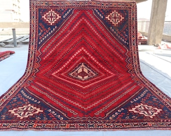 Puede incluir: Una alfombra grande, roja, azul y blanca con un diseño en forma de diamante en el centro. La alfombra tiene un borde con flecos.