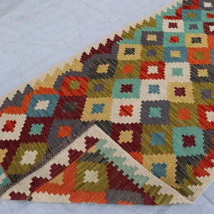 May include: A colorful geometric patterned rug with a cream background and fringe on the edges. The rug features a diamond pattern with shades of red, orange, yellow, green, blue, and gray.