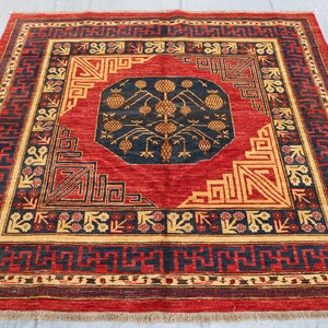 May include: A square, hand-woven rug with a central navy blue medallion featuring a tree-like design and golden accents. The rug has a red field with geometric patterns in gold and navy blue, and a dark red border.