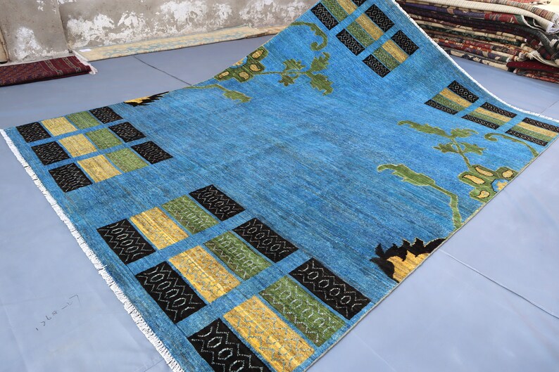 May include: A blue rug with a geometric pattern of black, yellow, and green squares. The rug has a fringe border and features a floral design in the center.