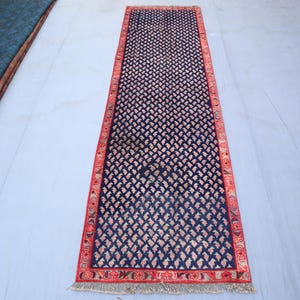 Peut inclure: Un tapis rectangulaire bleu marine avec un motif floral répétitif rose clair. Le tapis a une bordure rouge avec des motifs complexes et une bordure à franges. Le tapis est présenté sur une surface bleu clair.