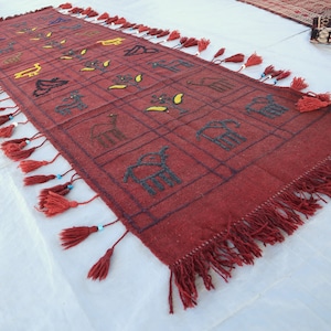 May include: A red woven rug with a geometric pattern of squares and rectangles. The rug has a fringe border and is decorated with embroidered animals and plants.