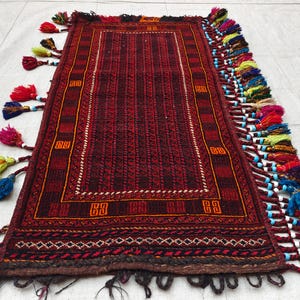 May include: A rectangular woven rug featuring a deep red and black geometric pattern with a central panel of vertical stripes. The edges are adorned with long, multi-colored yarn tassels in shades of red, blue, yellow, and green, accented with small white beads.