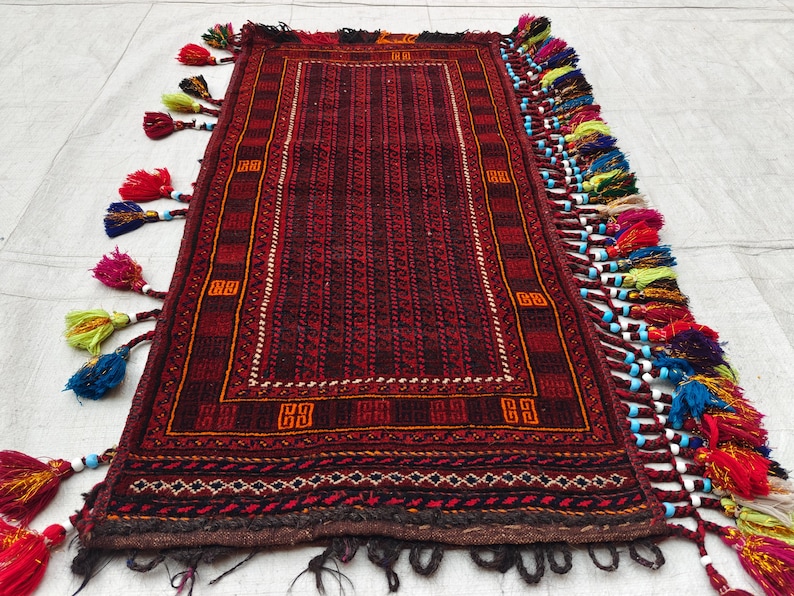 May include: A rectangular woven rug featuring a deep red and black geometric pattern with a central panel of vertical stripes. The edges are adorned with long, multi-colored yarn tassels in shades of red, blue, yellow, and green, accented with small white beads.