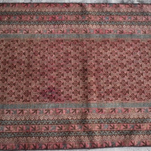 Pode incluir: Um tapete vintage com um padrão geométrico repetitivo em tons de marrom, rosa e verde. O tapete tem um aspecto desbotado e desgastado, o que aumenta o seu charme antigo.