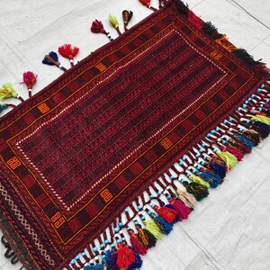 May include: A rectangular woven rug featuring a deep red and black geometric pattern with a central border. The edges are adorned with numerous multi-colored tassels in shades of blue, yellow, red, and green, accented with small white and blue beads.