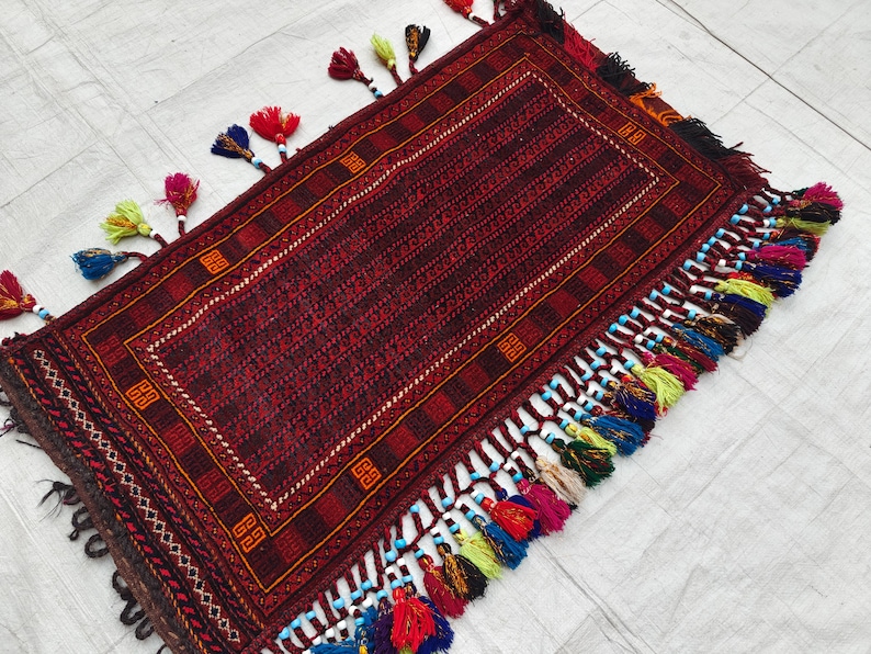 May include: A rectangular woven rug featuring a deep red and black geometric pattern with a central border. The edges are adorned with numerous multi-colored tassels in shades of blue, yellow, red, and green, accented with small white and blue beads.