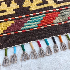 Op de afbeelding: Een geweven kleed met een donkerbruine basis en geometrische patronen in geel, turquoise, rood en grijs. De rand is geaccentueerd met een horizontale rode stiklijn en een rij veelkleurige kwastjes, elk met een omwikkelde bovenkant in diverse kleuren.