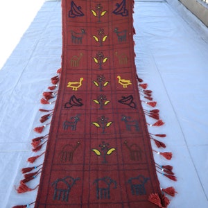 May include: A long, red rug with a geometric pattern of animals and plants. The rug has fringe on both ends.