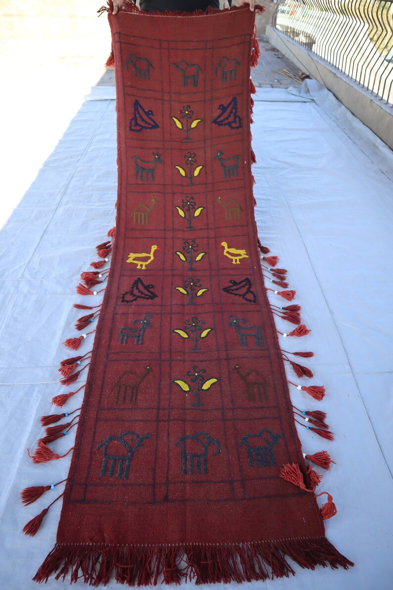May include: A long, red rug with a geometric pattern of animals and plants. The rug has fringe on both ends.