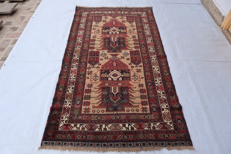 Pu&ograve; includere: Un tappeto tradizionale con sfondo beige e intricati motivi in rosso, nero e verde. Il tappeto presenta un disegno centrale con un motivo ripetitivo di alberi stilizzati e forme geometriche.