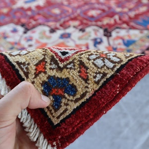 May include: Close-up of a red, cream, and blue patterned rug with fringe. The rug is folded in half and being held by a hand.