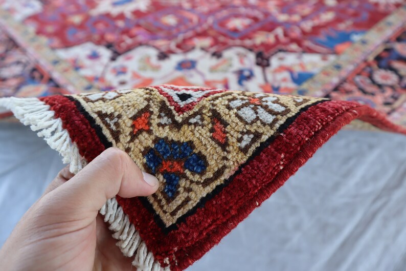 May include: Close-up of a red, cream, and blue patterned rug with fringe. The rug is folded in half and being held by a hand.