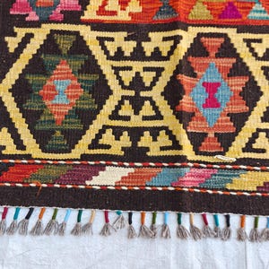 Op de afbeelding: Een geweven kleed met een donkerbruine achtergrond en herhalende geometrische ruitpatronen in geel, oranje, rood, blauw en groen. De onderrand toont een veelkleurige zigzagrand en een rij korte, lichtgrijze franjes aan de onderzijde.