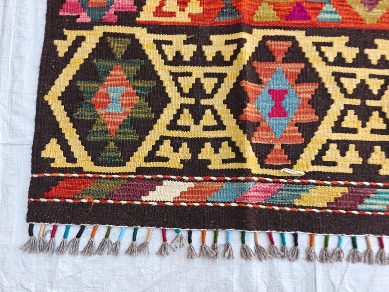 Op de afbeelding: Een geweven kleed met een donkerbruine achtergrond en herhalende geometrische ruitpatronen in geel, oranje, rood, blauw en groen. De onderrand toont een veelkleurige zigzagrand en een rij korte, lichtgrijze franjes aan de onderzijde.