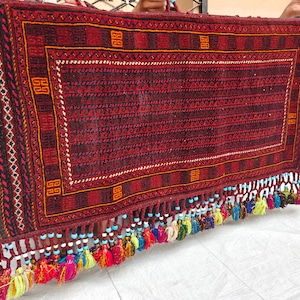 May include: A rectangular woven textile featuring a deep red and black geometric pattern with a central field of repeating wave motifs. The bottom edge is adorned with a row of multi-colored tassels, each accented with small light blue and white beads.