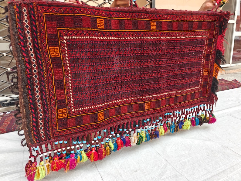 May include: A rectangular woven textile featuring a deep red and black geometric pattern with a central field of repeating wave motifs. The bottom edge is adorned with a row of multi-colored tassels, each accented with small light blue and white beads.