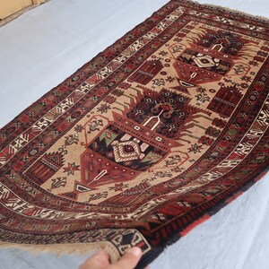 Pu&ograve; includere: Un tappeto tradizionale con sfondo beige e intricati motivi in rosso, nero e verde. Il tappeto presenta un disegno centrale di due alberi stilizzati con motivi floreali.
