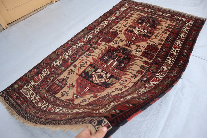 Pu&ograve; includere: Un tappeto tradizionale con sfondo beige e intricati motivi in rosso, nero e verde. Il tappeto presenta un disegno centrale di due alberi stilizzati con motivi floreali.